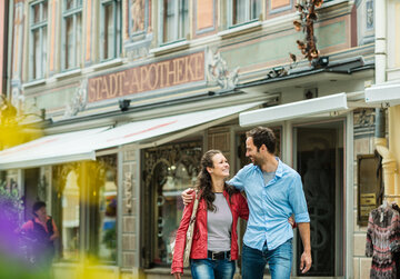Das Bild zeigt einen jungen Mann und eine Frau. Der Mann hat den Arm um die Frau gelegt. Sie lächeln sich an. Die beiden stehen vor einem reich verzierten Gebäude. Im Hintergrund sind Schaufenster zu sehen. Es ist Sommer. 