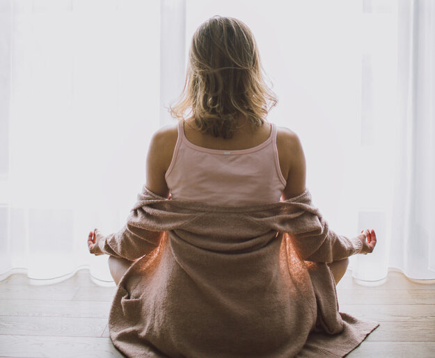 Das Bild zeigt eine Frau, die im Schneidersitz auf dem Holzboden sitzt. Sie sitzt in Richtung eines hellen Fensters, das mit einem bodenlangen Stohr verhängt ist.  