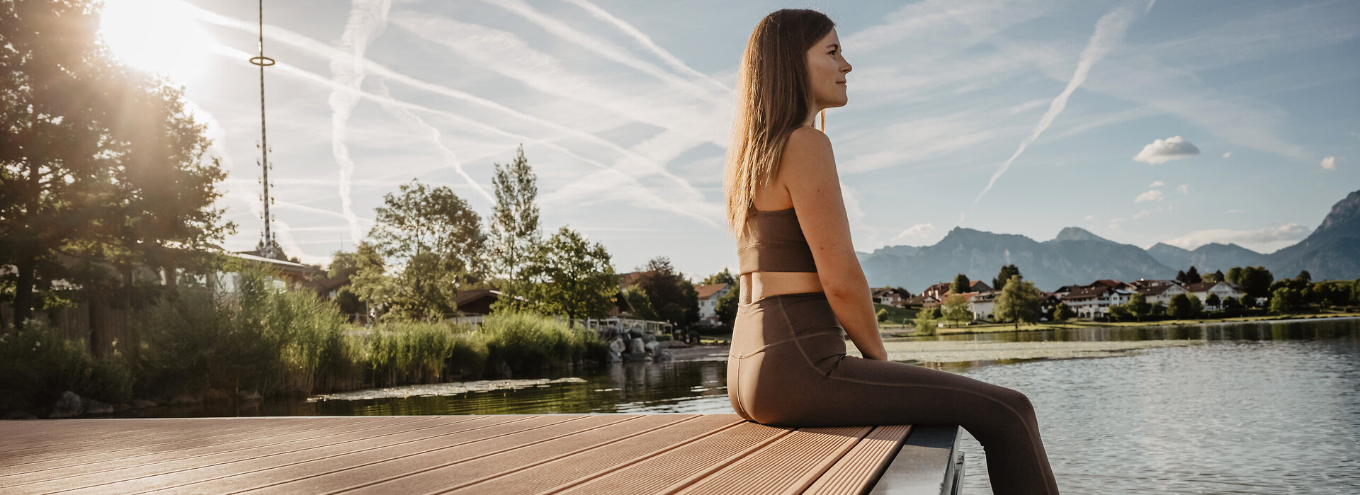 Das Bild zeigt eine Frau, die auf einem Steg am Hopfensee mit Blick auf die Berge sitzt