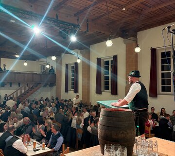 Hier hält ein Mann die begrüßungsrede des Starkbierabends. Auf einem Tisch steht ein Starkbierfass welches zum Anstich bereit steht.