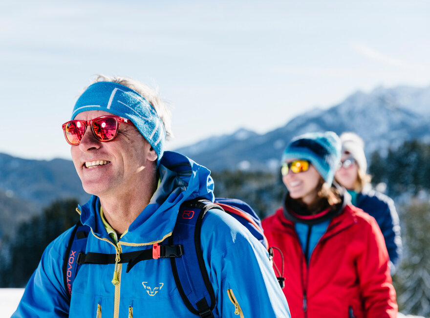 Das Bild zeigt drei Personen in sportlicher Winterkleidung mit Sonnenbrillen und Mützen. Sie laufen hintereinander. Im Hintergrund erheben sich bewaldete, schneebedeckte Berge. Der Himmel ist blau. Die Sonne scheint. Ein Mann läuft voraus. Zwei Frau laufen ihm nach. Alle lächeln. Man kann nur ihre Oberköper und Köpfe sehen. 