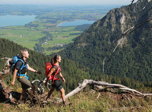 Das Bild zeigt einen Mann und eine Frau beim Wandern in den Bergen. Sie tragen kurze Wanderkleidung. Sie haben bereits einige Höhenmeter zurückgelegt. Im Tal sind grüne Wiesen, Wälder und Seen zu sehen. Die Sonne scheint.