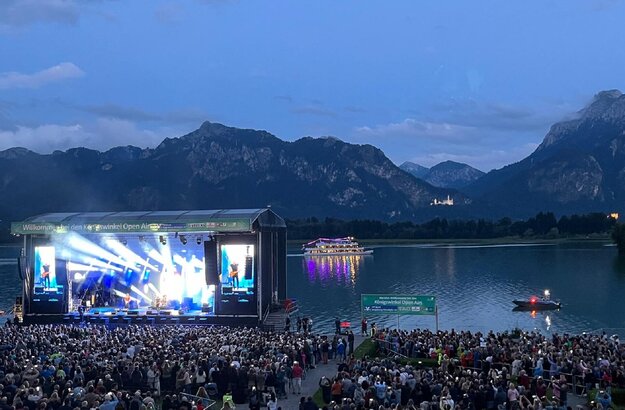 Das Foto zeigt eine große, hell erleuchtete Open Air Bühne am Ufer des Forggensees. Im Vordergrund stehen unzählige Personen. Im Hintergrund sind die Berge und das beleuchtete Schloss Neuschwanstein zu sehen, davor liegt der Forggensee auf dem ein beleuchtetes Schiff fährt.  