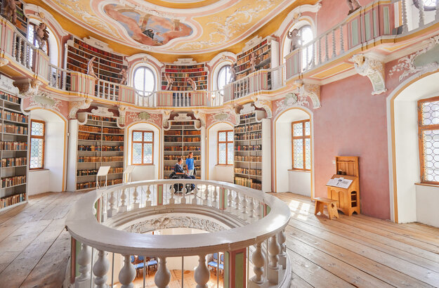 Die Bibliothek des Hohen Schlosses in Füssen mit hohen Regalen und einem beeindruckenden Rundbogen. Das Bild zeigt einen Raum voller Wissen und Geschichte.