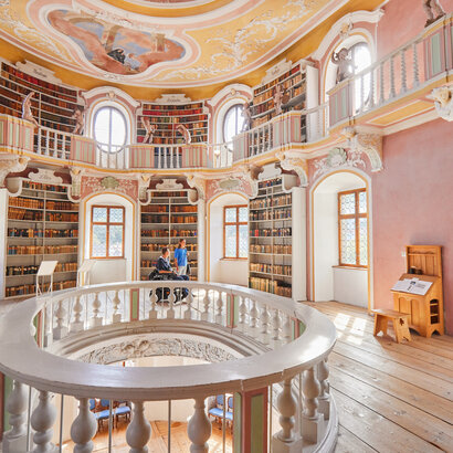 Die Bibliothek des Hohen Schlosses in Füssen mit hohen Regalen und einem beeindruckenden Rundbogen. Das Bild zeigt einen Raum voller Wissen und Geschichte.