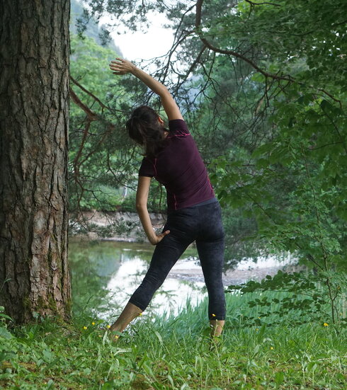 Das Bild zeigt eine Frau in sportlichem Outfit mit dunklen Haaren, die eine Yoga-Bewegung macht. Sie steht in der Natur unter einem Baum im hohen Gras und blickt auf ein Gewässer.