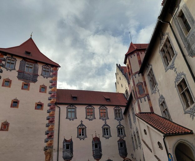 Illusionsmalereien am Hohen Schloss in Füssen Das Bild zeigt ein großes, dekoratives Gebäude mit einer reichen architektonischen Geschichte. Das Gebäude ist in einem hellen Beige-Ton gestrichen, mit Akzenten von Rot und Braun, die Tiefe und Kontrast hinzufügen. Das Dach des Gebäudes ist mit mehreren kleinen Fenstern verziert, und ein großes, spitzes Dach krönt die Struktur. Die Fassade des Gebäudes ist mit komplexen Details und Mustern verziert, die das Handwerk einer vergangenen Ära widerspiegeln.