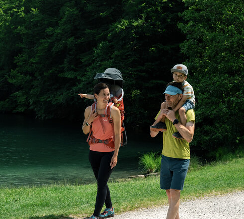 Eine Familie wandert auf einem Weg entlang eines Gewässers. Die Mutter trägt ein Kind im Tragegurt, während der Vater einen anderen Jungen auf den Schultern hat. Die Umgebung ist grün und natürliche, mit Bäumen im Hintergrund. Es ist ein sonniger Tag.