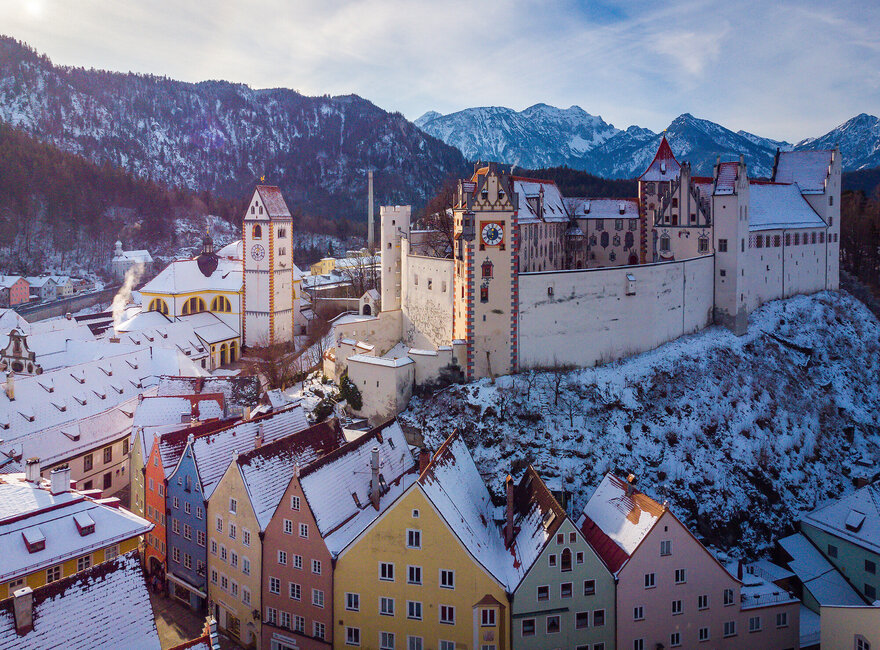 Winterliche Ansicht der Füssener Altstadt mit schneebedeckten Dächern, farbenfrohen Gebäuden, einer Kirche mit Turm, und dem Hohen Schloss vor einer Bergkette.
