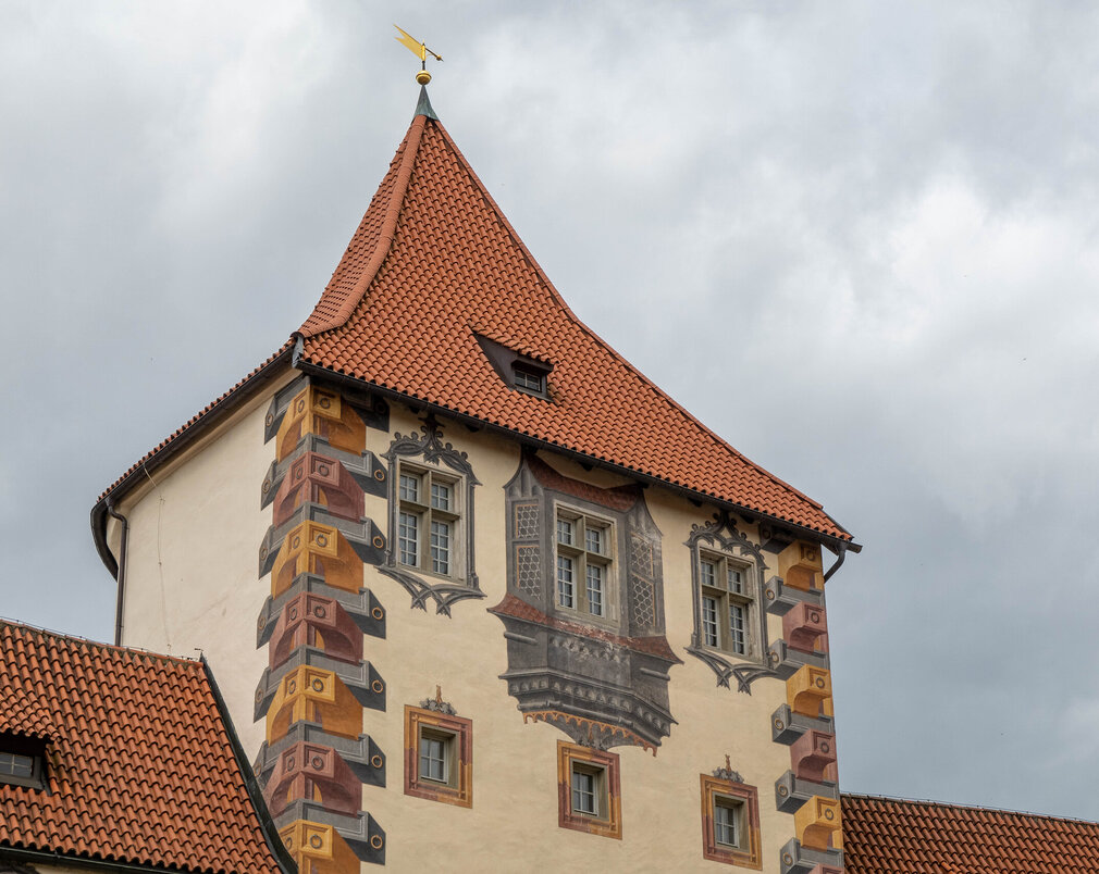 Das Bild zeigt ein großes, reich verziertes Gebäude mit einem charakteristischen Architekturstil. Das Gebäude ist in einer Vielzahl von Farben gestaltet, darunter Töne von Gelb, Orange und Braun. Das Dach des Gebäudes ist rot und weist eine spitze, konische Form auf. Auf dem Dach ist eine kleine gelbe Fahne. Das Gebäude hat mehrere Fenster, die alle mit filigranen Designs und Mustern verziert sind. Der Hintergrund ist wolkig, was auf einen bedeckten Tag hindeutet. Der Eindruck ist der eines großen, historischen Gebäudes, möglicherweise eines Schlosses oder einer Kirche, mit einem reichen und detaillierten architektonischen Design.