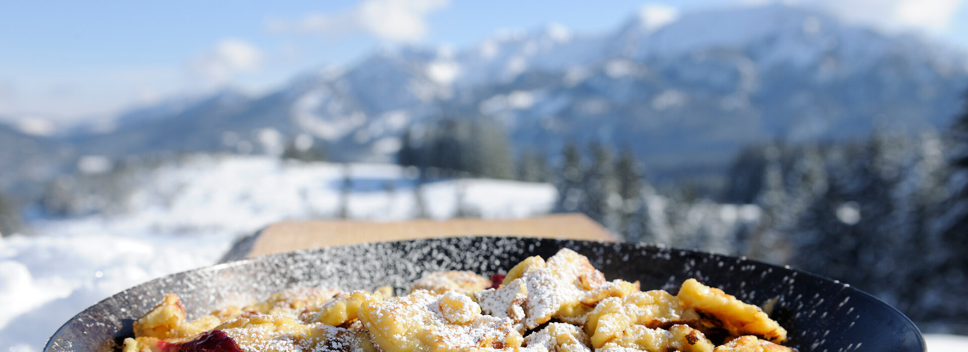 Eine Pfanne mit Kaiserschmarren mit Puderzucker im Vordergrund vor einer verschneiten Bergkulisse. 