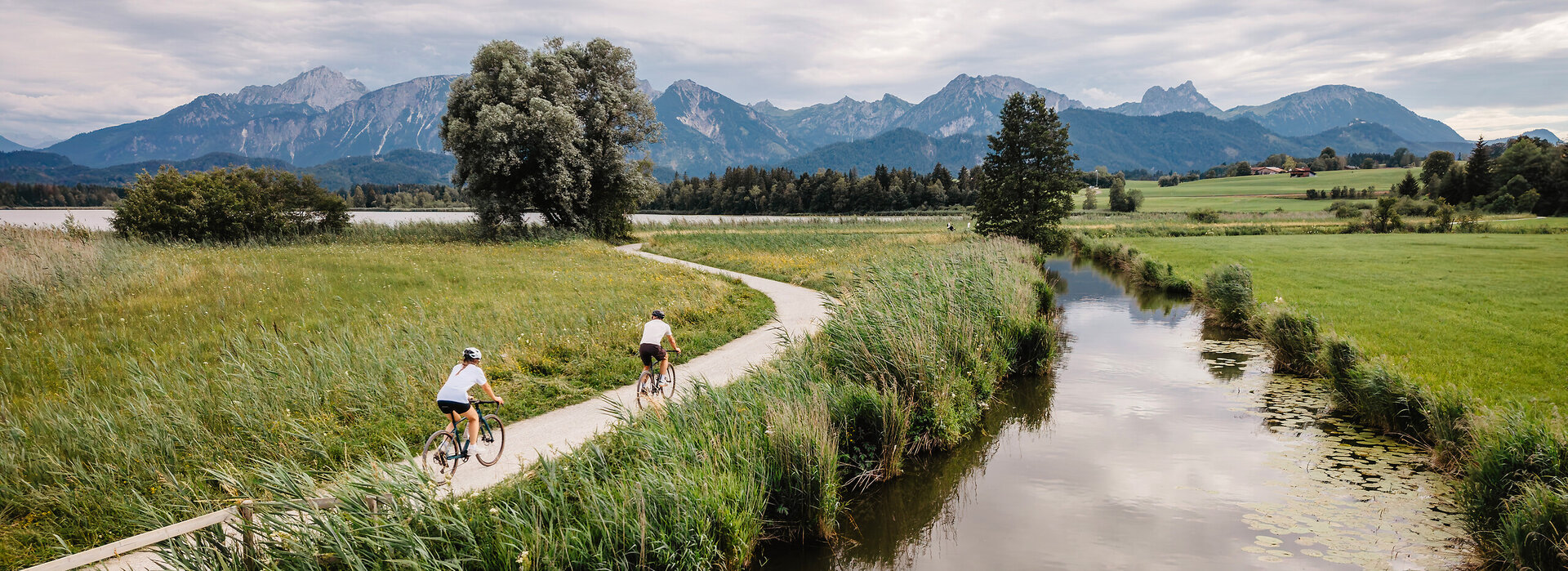 Das Bild zeigt einen dunkel schimmerenden Bachlauf entlang eines Schilfgürtels, durch den ein breiter Kiesweg führt. Auf dem Weg sind zwei Fahrradfahrende zu erkennen. Im Hintergrund erheben sich Berge.