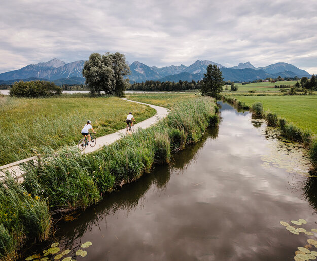 Das Bild zeigt einen dunkel schimmerenden Bachlauf entlang eines Schilfgürtels, durch den ein breiter Kiesweg führt. Auf dem Weg sind zwei Fahrradfahrende zu erkennen. Im Hintergrund erheben sich Berge.
