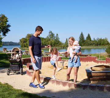 ie Szenene in dem Bild ist eine friedliche Momentaufnahme in einem Park. Vor einem ruhigen See stehen ein Mann und eine Frau, beide tragen legeres Outfit. Der Mann hat ein blaues T-Shirt und blaue Shorts an, während die Frau ein weißes T-Shirt und blaue Shorts trägt. Sie halten Händchen, was ihre Nähe zum Ausdruck bringt.  Im Hintergrund ist ein friedlicher Teich zu sehen, das zur friedlichen Atmosphäre des Parks beiträgt. Einige Bäume stehen dort und bieten eine natürliche Kulisse für die Szene.  Der Park ist auch mit einem Spielplatz ausgestattet, inklusive eines Sandkastens und eines Schaukelsatzes. Der Sandkasten liegt nahe zum See, bietet also eine fröhliche und aktive Spielmöglichkeit für Kinder. Der Schaukelsatz befindet sich etwas weiter weg, wo Kinder ungetehrt spielen und Eltern ihnen zuschauen können.  Das Bild zeigt generell einen typischen Tag im Park, mit Leuten, die die Natur genießen, und Kindern, die im Sandkasten und auf den Schaukeln spielen.