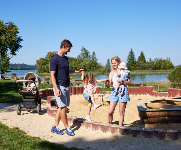 ie Szenene in dem Bild ist eine friedliche Momentaufnahme in einem Park. Vor einem ruhigen See stehen ein Mann und eine Frau, beide tragen legeres Outfit. Der Mann hat ein blaues T-Shirt und blaue Shorts an, während die Frau ein weißes T-Shirt und blaue Shorts trägt. Sie halten Händchen, was ihre Nähe zum Ausdruck bringt.  Im Hintergrund ist ein friedlicher Teich zu sehen, das zur friedlichen Atmosphäre des Parks beiträgt. Einige Bäume stehen dort und bieten eine natürliche Kulisse für die Szene.  Der Park ist auch mit einem Spielplatz ausgestattet, inklusive eines Sandkastens und eines Schaukelsatzes. Der Sandkasten liegt nahe zum See, bietet also eine fröhliche und aktive Spielmöglichkeit für Kinder. Der Schaukelsatz befindet sich etwas weiter weg, wo Kinder ungetehrt spielen und Eltern ihnen zuschauen können.  Das Bild zeigt generell einen typischen Tag im Park, mit Leuten, die die Natur genießen, und Kindern, die im Sandkasten und auf den Schaukeln spielen.