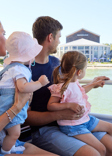 Das Bild zeigt einen friedlichen Moment auf einem Boot. Eine Familie von vier Personen sitzt zusammen und genießt die Aussicht auf das Wasser. Die Mutter, in einem weißen Bluse gekleidet, hält ihr Baby auf dem Arm. Der Vater trägt ein blaues Hemd und hält ihre Tochter auf dem Schoß. Die Tochter ist in einem rosa Shirt gekleidet und sitzt auf dem Schoß des Vaters. Das Boot, auf dem sie sind, ist weiß und verfügt über eine Reling. Im Hintergrund ist ein Gebäude und Bäume zu sehen, die zur malerischen Szene beitragen. Die Familie scheint einen großartigen Tag zusammen zu verbringen und genießt ihre Bootsfahrt voll und ganz.