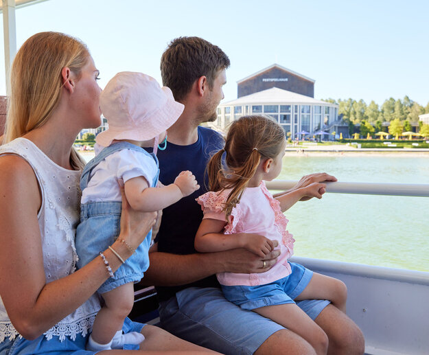 Das Bild zeigt einen friedlichen Moment auf einem Boot. Eine Familie von vier Personen sitzt zusammen und genießt die Aussicht auf das Wasser. Die Mutter, in einem weißen Bluse gekleidet, hält ihr Baby auf dem Arm. Der Vater trägt ein blaues Hemd und hält ihre Tochter auf dem Schoß. Die Tochter ist in einem rosa Shirt gekleidet und sitzt auf dem Schoß des Vaters. Das Boot, auf dem sie sind, ist weiß und verfügt über eine Reling. Im Hintergrund ist ein Gebäude und Bäume zu sehen, die zur malerischen Szene beitragen. Die Familie scheint einen großartigen Tag zusammen zu verbringen und genießt ihre Bootsfahrt voll und ganz.