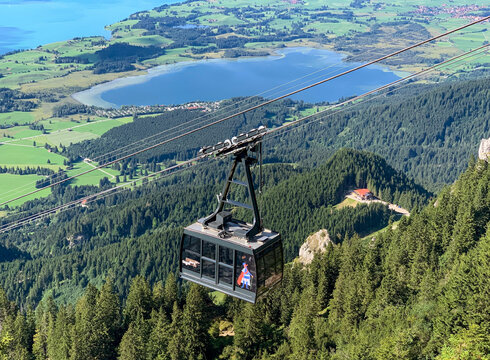 Das Bild zeigt die an einem Seil hängende Kabine einer Bergbahn. Im Tal erblickt man zwei blaue Seen, Wiesen und Wälder. Die Sonne scheint. 