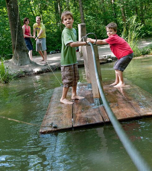 Das Bild zeigt zwei Jungen, die auf einem hölzernen Floß über einen Bach fahren. Dabei halten sie sich an einem Stahlseil fest, das über den Bach gespannt ist. Im Hintergrund stehen ein Mann und eine Frau und lenken das Floß. Ringsum wachsen hohe Bäume. Die Sonne scheint. Das Wasser des Bachs ist trüb. Die Kinder haben sichtlich Spaß.