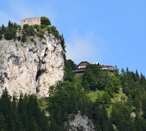 Auf dem Bild ist ein steiler, bewaldeter Hang mit einem Felsen zu sehen. Am oberen Ende der Felsen steht eine Burgruine, während weiter unten ein modernes Gebäude inmitten von Bäumen sichtbar ist. Der Himmel ist blau mit wenigen Wolken.