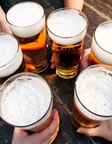 hier erkennt man fünf Hände, die mit vollen Biergläsern die auf einem Holztisch  anstoßen. 