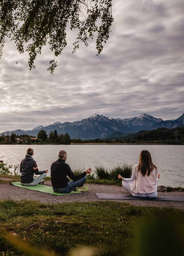 Das Bild zeigt drei Menschen, die am Seeufer sitzen und meditieren