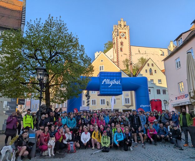 Das Bild zeigt eine große Ansammlung von Menschen in Wanderbekleidung in der Füssener Fußgängerzone. Im Hintergrund stehen ein großer Allgäu-Torbogen und das Hohe Schloss von Füssen strahlt in der Morgensonne.