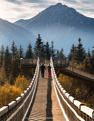 Zwei Personen spazieren über den Baumkronenweg im Walderlebniszentrum in Füssen. Die Brücke bietet einen atemberaubenden Blick auf die umliegenden Berge.