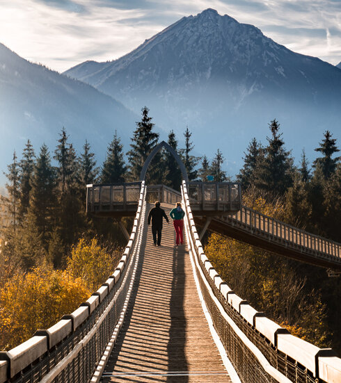 Zwei Personen spazieren über den Baumkronenweg im Walderlebniszentrum in Füssen. Die Brücke bietet einen atemberaubenden Blick auf die umliegenden Berge.