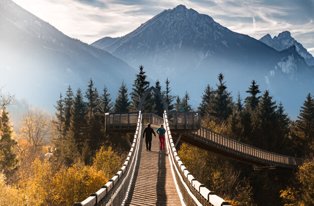 Zwei Personen spazieren über den Baumkronenweg im Walderlebniszentrum in Füssen. Die Brücke bietet einen atemberaubenden Blick auf die umliegenden Berge.