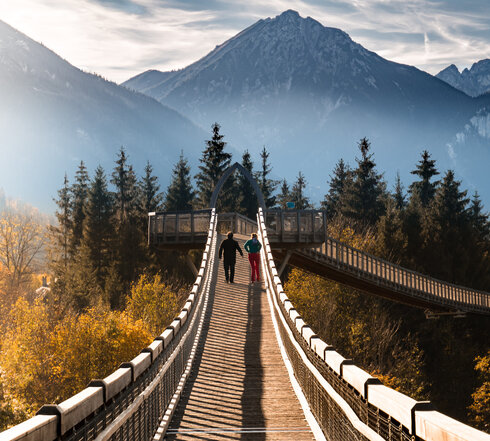 Zwei Personen spazieren über den Baumkronenweg im Walderlebniszentrum in Füssen. Die Brücke bietet einen atemberaubenden Blick auf die umliegenden Berge.