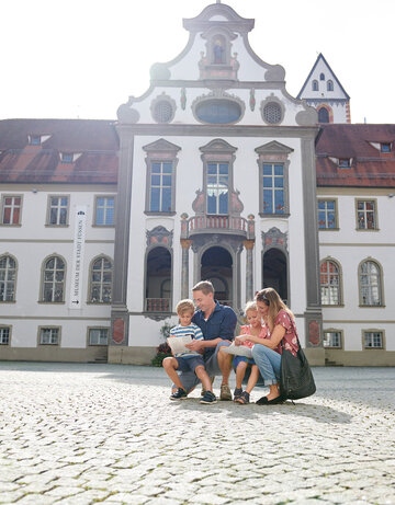 Die Familie sitzt auf einem Steinpflaster vor einem großen, historischen Gebäude. Das Gebäude ist weiß und hat eine rote Dachterrasse. Es hat viele Fenster und ist mit einem großen Turm versehen. Die Familie besteht aus einem Mann, einer Frau und zwei Kindern. Der Mann und die Frau sind beide in blauen T-Shirts. Die Frau hält ein Kind auf ihrem Schoß. Die Familie scheint eine Pause zu machen, während sie auf dem Steinpflaster sitzen.
