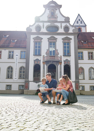 Die Familie sitzt auf einem Steinpflaster vor einem großen, historischen Gebäude. Das Gebäude ist weiß und hat eine rote Dachterrasse. Es hat viele Fenster und ist mit einem großen Turm versehen. Die Familie besteht aus einem Mann, einer Frau und zwei Kindern. Der Mann und die Frau sind beide in blauen T-Shirts. Die Frau hält ein Kind auf ihrem Schoß. Die Familie scheint eine Pause zu machen, während sie auf dem Steinpflaster sitzen.