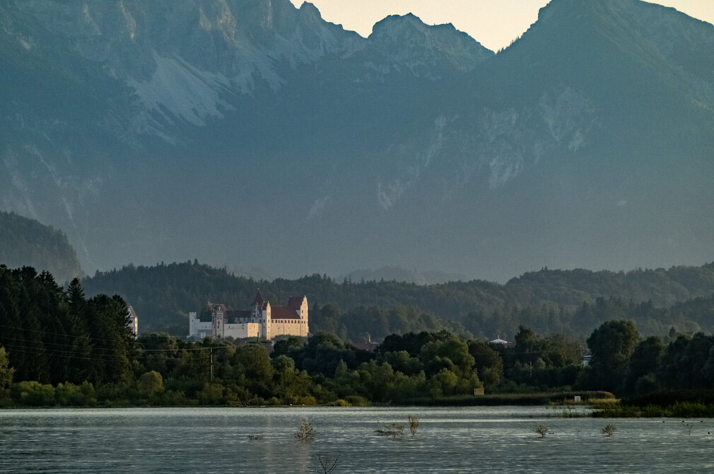 Das Bild zeigt eine friedliche Landschaft, die von einem majestätischen Schloss dominiert wird. Es handelt sich hier um das Hohe Schloss bei Füssen, das mit seinen weißen Wänden und dem roten Dach vor dem Bergpanorama steht. 