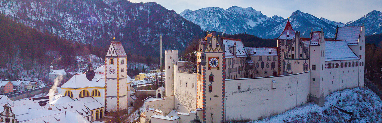 Das Bild fängt die atemberaubende Sicht auf das mittelalterliche Städtchen Füssen in Deutschland ein. Die Stadt liegt auf einem Hügel, von majestätischen Bergen umgeben. Die Architektur der Stadt ist durch bunte Gebäude geprägt, jedes mit einem einzigartigen Charme. Die Gebäude sind mit Schnee verziert, was eine Prise Winter in die Szene bringt