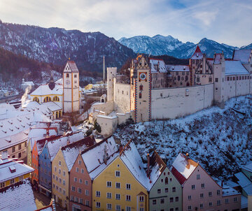 Das Bild fängt die atemberaubende Sicht auf das mittelalterliche Städtchen Füssen in Deutschland ein. Die Stadt liegt auf einem Hügel, von majestätischen Bergen umgeben. Die Architektur der Stadt ist durch bunte Gebäude geprägt, jedes mit einem einzigartigen Charme. Die Gebäude sind mit Schnee verziert, was eine Prise Winter in die Szene bringt