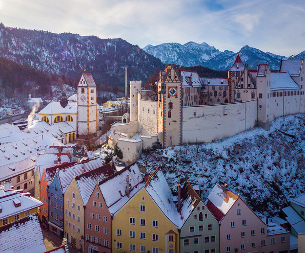 Das Bild fängt die atemberaubende Sicht auf das mittelalterliche Städtchen Füssen in Deutschland ein. Die Stadt liegt auf einem Hügel, von majestätischen Bergen umgeben. Die Architektur der Stadt ist durch bunte Gebäude geprägt, jedes mit einem einzigartigen Charme. Die Gebäude sind mit Schnee verziert, was eine Prise Winter in die Szene bringt