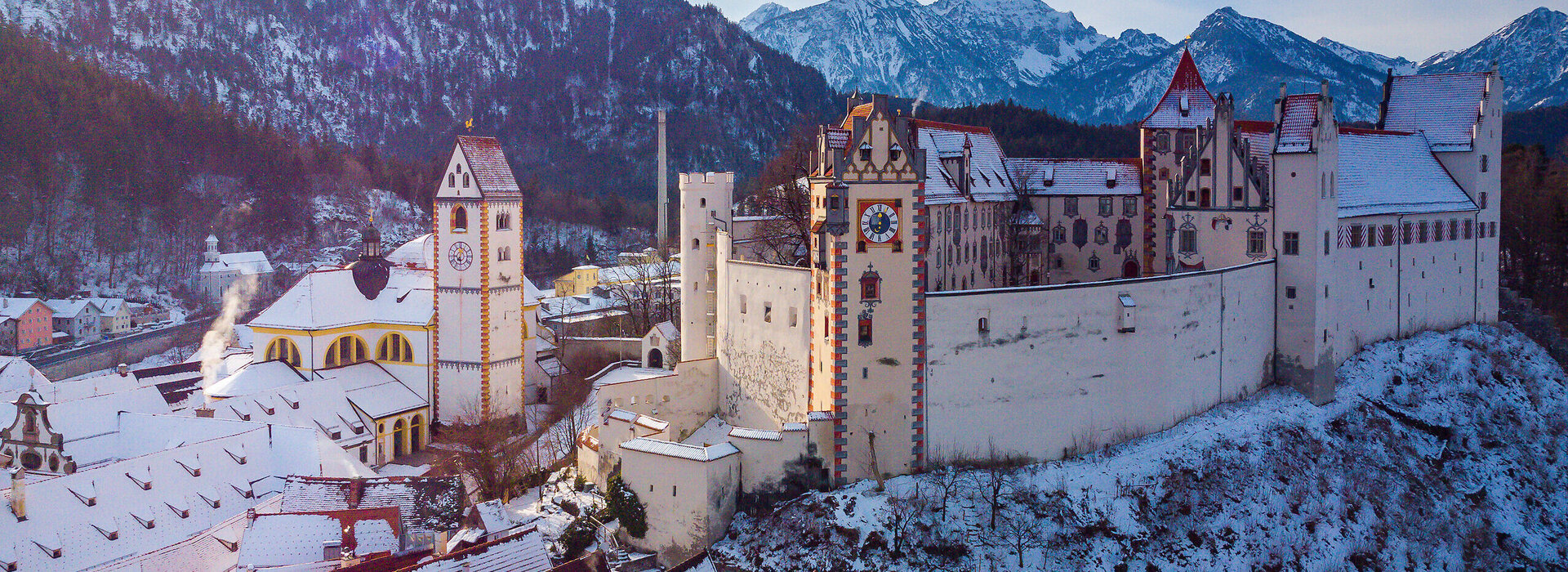 The picture captures the breathtaking view of the medieval town of Füssen in Germany. The town is situated on a hill surrounded by majestic mountains. The architecture of the town is characterized by colourful buildings, each with a unique charm. The buildings are decorated with snow, adding a touch of winter to the scene