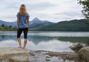 Das Bild zeigt eine blonde Frau, die auf einem großen Stein am Ufer eines Sees steht. Sie dreht dem Betrachter den Rücken zu. Sie trägt eine kurze Hose und ein T-Shirt. Der See ist von grünen Wäldern umgeben. Im Hintergrund erheben sich hohe Berge. Der Himmel ist wolkenverhangen. 