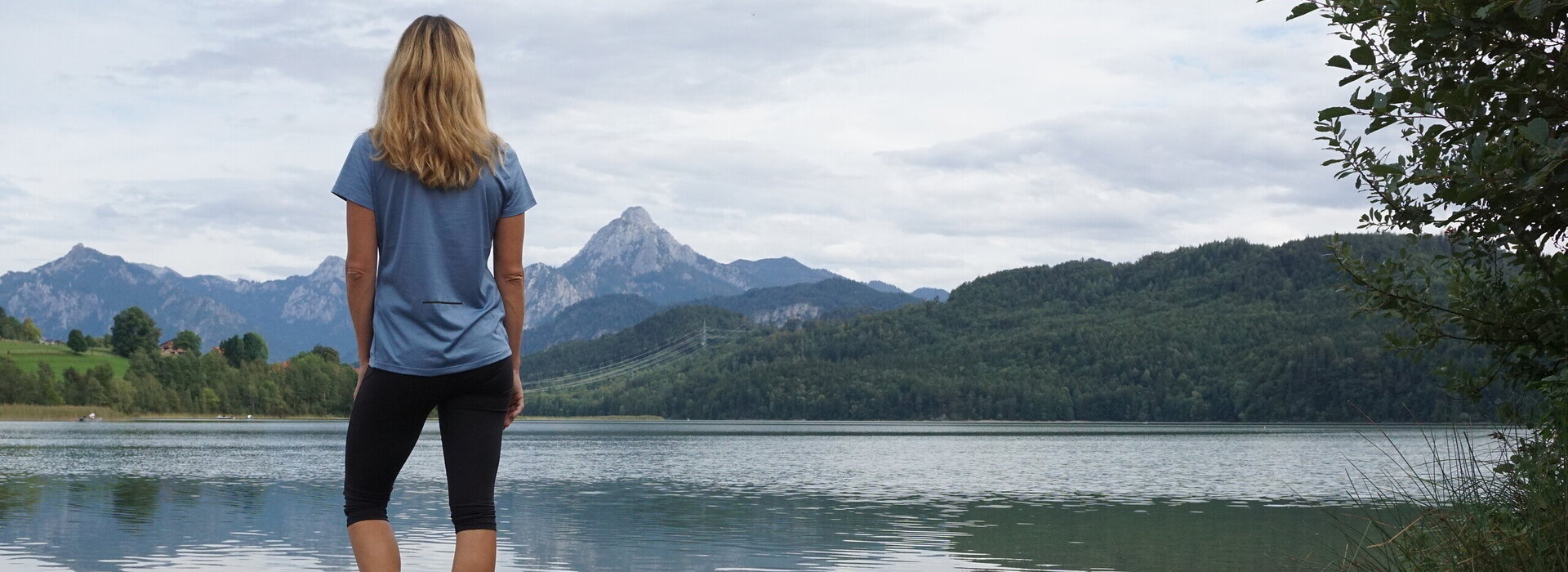 Das Bild zeigt eine blonde Frau, die auf einem großen Stein am Ufer eines Sees steht. Sie dreht dem Betrachter den Rücken zu. Sie trägt eine kurze Hose und ein T-Shirt. Der See ist von grünen Wäldern umgeben. Im Hintergrund erheben sich hohe Berge. Der Himmel ist wolkenverhangen. 
