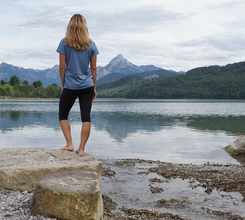 Das Bild zeigt eine blonde Frau, die auf einem großen Stein am Ufer eines Sees steht. Sie dreht dem Betrachter den Rücken zu. Sie trägt eine kurze Hose und ein T-Shirt. Der See ist von grünen Wäldern umgeben. Im Hintergrund erheben sich hohe Berge. Der Himmel ist wolkenverhangen. 