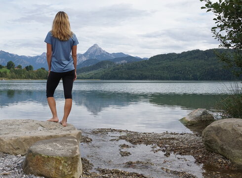 Das Bild zeigt eine blonde Frau, die auf einem großen Stein am Ufer eines Sees steht. Sie dreht dem Betrachter den Rücken zu. Sie trägt eine kurze Hose und ein T-Shirt. Der See ist von grünen Wäldern umgeben. Im Hintergrund erheben sich hohe Berge. Der Himmel ist wolkenverhangen. 
