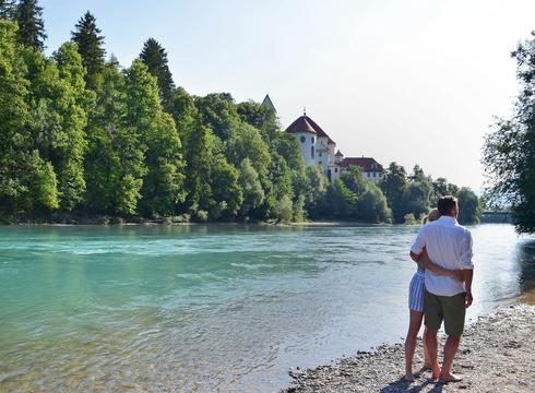 Das Bild zeigt ein Paar, das an einem steinigen Ufer des Flusses Lech steht. Der Mann und die Frau stehen dicht beieinander, ihre Körper sind dem Wasser zugewandt. Der türkise Fluss liegt im Fokus des Bildes. Auf der gegenüberliegenden Seite erhebt sich ein großer Gebäudekomplex mit weißen Mauern und einem roten Dach. Hierbei handelt es sich um das Hohe Schloss und das ehemalige Kloster St. Mang in Füssen. Der Himmel darüber ist klar, blau und wolkenlos.