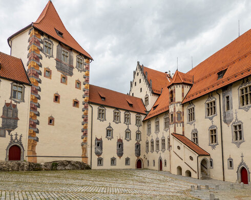 Das Bild zeigt den Innenhof des Hohen Schlosses in Füssen, der durch seine Illusionsmalerei an den Fassaden beeindruckt. Das Schlossgebäude hat rote Türen und ein Ziegeldach.