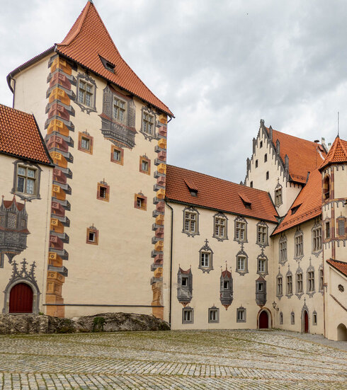 Das Bild zeigt den Innenhof des Hohen Schlosses in Füssen, der durch seine Illusionsmalerei an den Fassaden beeindruckt. Das Schlossgebäude hat rote Türen und ein Ziegeldach.