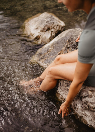 Das Bild zeigt eine Frau, die ihre Füße in das Wasser eines Sees baumeln lässt. Sie sitzt auf großen Steinen, die im Wasser liegen.