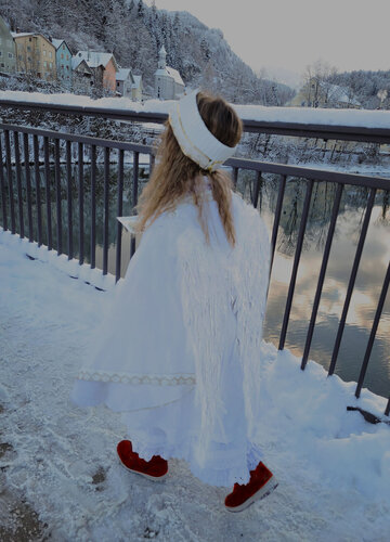 Das Bild fängt eine idyllische Winterlandschaft ein. Eine junge Frau, angekleidet in ein weißes Kleid und einen passenden weißen Hut, ist die Hauptperson des Fotos. Sie ist dabei, auf einem schneebedeckten Bürgersteig zu gehen, ihre roten Schuhe kontrastieren mit dem weißen Schnee. Die Frau sieht nach unten, vielleicht auf den Schnee unter ihren Füßen.  Im Hintergrund gibt es eine schöne Aussicht auf einen Fluss, über den eine Brücke spannt. Die Brücke ist mit Schnee verziert, was zur wintersportlichen Atmosphäre hinzufügt. Auf der anderen Seite des Flusses gibt es bunte Häuser, deren leuchtende Farben sich gegen den weißen Schnee abheben.  Der Himmel über ist bedeckt, was ein sanftes Licht über die gesamte Szene wirft. Das Bild ist eine wunderschöne Darstellung eines Wintertages, mit der Reise der Frau entlang des schneebedeckten Bürgersteiges als Höhepunkt