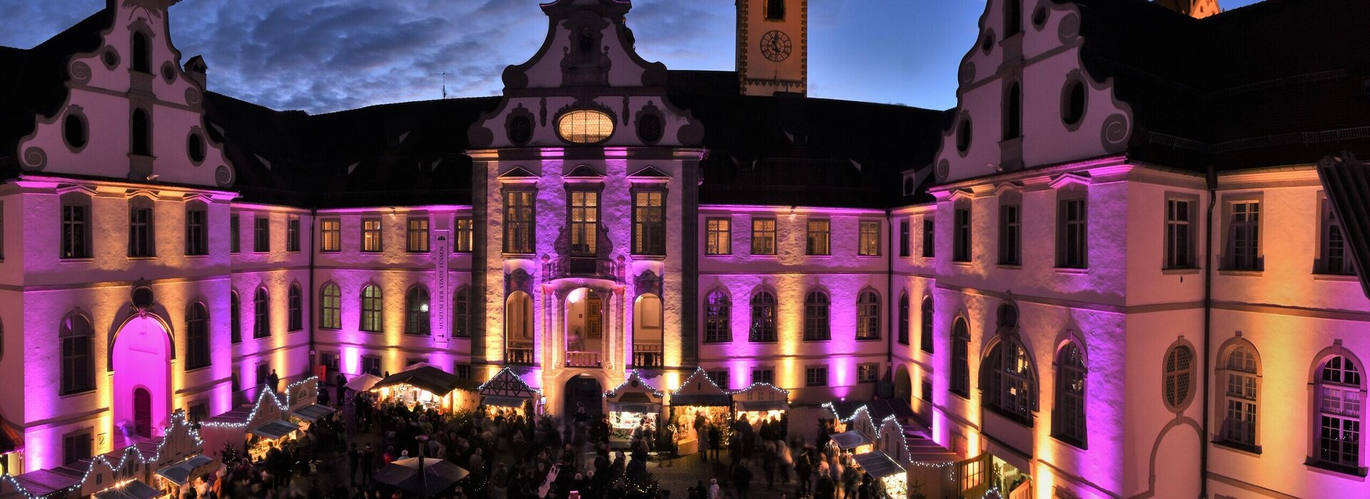 Das Bild zeigt den Füssener Adventsmarkt im Klosterhof des ehemaligen Benediktinerklosters St. Mang. Die Fassade des rundumlaufenden Gebäudes ist pink beleuchtet. Die Buden des Adventsmarkt sind ebenfalls festlich beleuchtet. Zahlreiche Menschen sind zu sehen. Gerade ist die Sonne untergegangen. Man sieht noch die letzten Sonnenstrahlen über dem Klosterdach.  