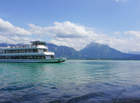 Das Bild fängt eine friedliche Szene eines weißen Schiffes mit drei Aussichtsdecks ein, das auf einem ruhigen blauen See – dem Forggensee - schwimmt. Das Schiff steht im Mittelpunkt des Bildes und wird von der unendlichen Weite des Forggensees umgeben. Der See selbst ist inmitten einer malerischen Kulisse von majestätischen Bergen eingebettet. Der klare und blaue Himmel ist vereinzelt von flauschigen, weißen Wolken durchzogen. 