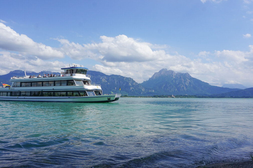 Das Bild fängt eine friedliche Szene eines weißen Schiffes mit drei Aussichtsdecks ein, das auf einem ruhigen blauen See – dem Forggensee - schwimmt. Das Schiff steht im Mittelpunkt des Bildes und wird von der unendlichen Weite des Forggensees umgeben. Der See selbst ist inmitten einer malerischen Kulisse von majestätischen Bergen eingebettet. Der klare und blaue Himmel ist vereinzelt von flauschigen, weißen Wolken durchzogen.